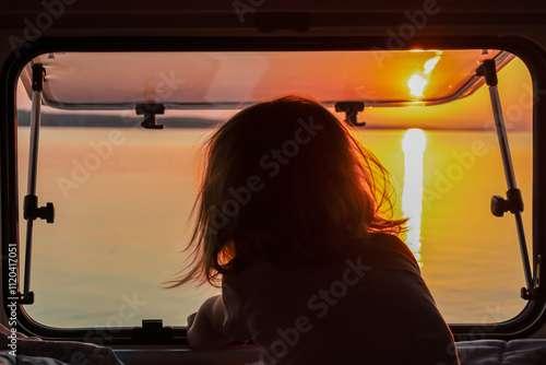 Child looking out of camper window with breathtaking fiery sunset over calm Adriatic Mediterranean Sea in Premantura, Istria, Croatia. Sun reflection on water creates shimmering path towards horizon photo