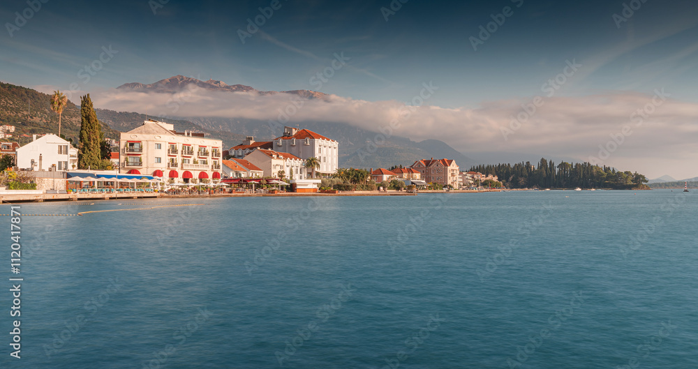 Scenic view of Tivat coastline featuring hotels, restaurants, and a small, verdant island under a misty sky Scenic view of Tivat coastline featuring hotels, restaurants, and a small, verdant island under a misty sky