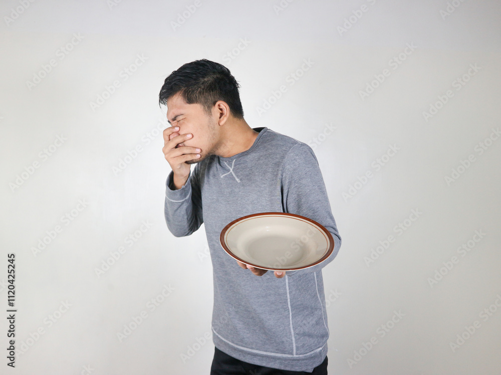 Asian man holding an empty plate and showing his distaste for food while covering his mouth. gesture of wanting to vomit Asian man holding an empty plate and showing his distaste for food while covering his mouth. gesture of wanting to vomit