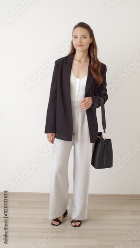 Young beautiful woman with long brunette hair, wearing black jacket, light grey pants, black bag and sandals, shows her outfit. Women clothes. Girl shows her outfit at home against a white wall.