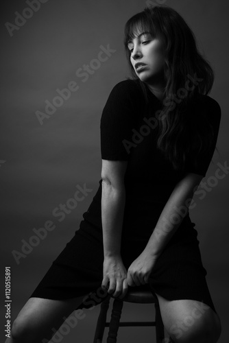 Black and white studio portrait of a caucasian woman with long dark hair with bangs. She is posed on a wooden stool wearing a black knit dress. She is looking to the left. 