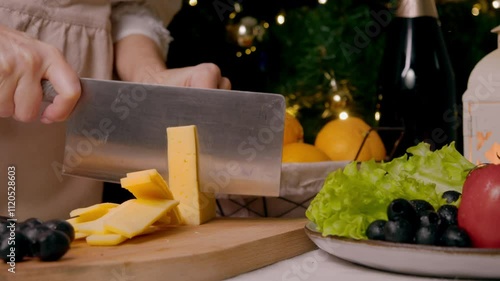 Cook cuts cheese into thin slices in order to serve it at Christmas table, which also includes variety of vegetables, fruits, and champagne. Close up