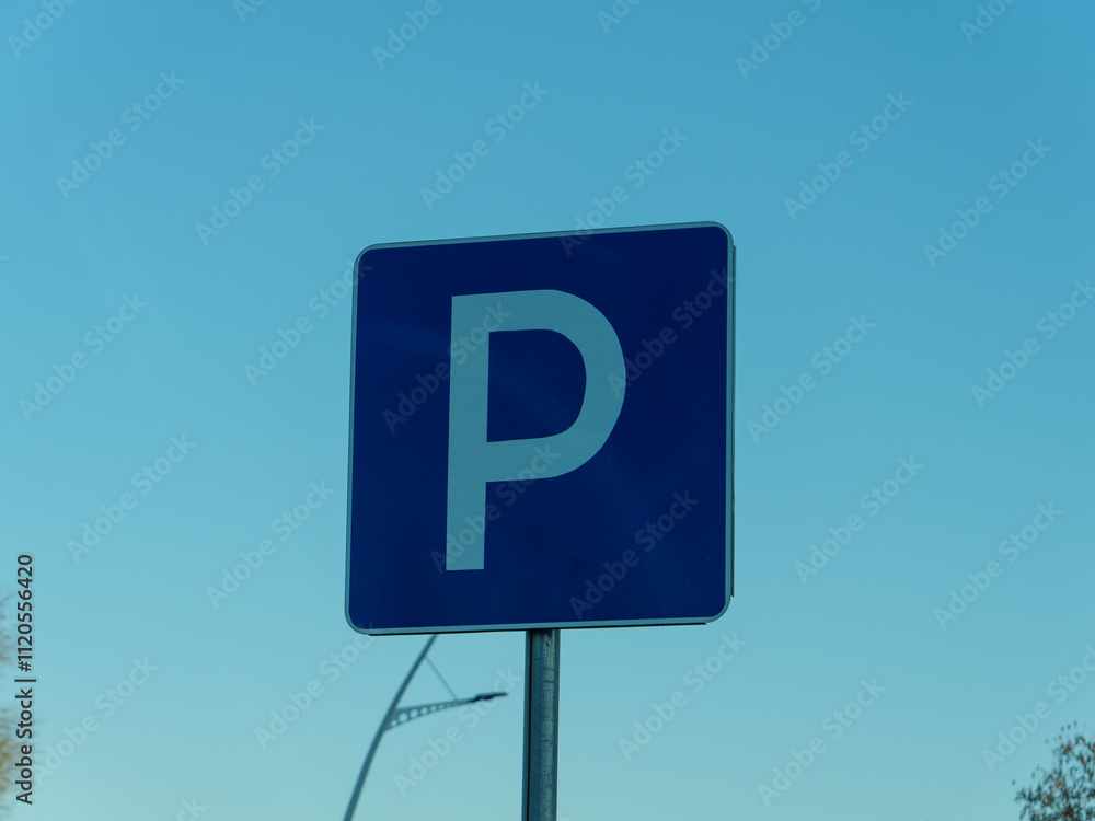 A blue parking sign stands tall against a clear sky, indicating a designated parking area close to a busy road. The surroundings are typical of an urban environment. A blue parking sign stands tall against a clear sky, indicating a designated parking area close to a busy road. The surroundings are typical of an urban environment.