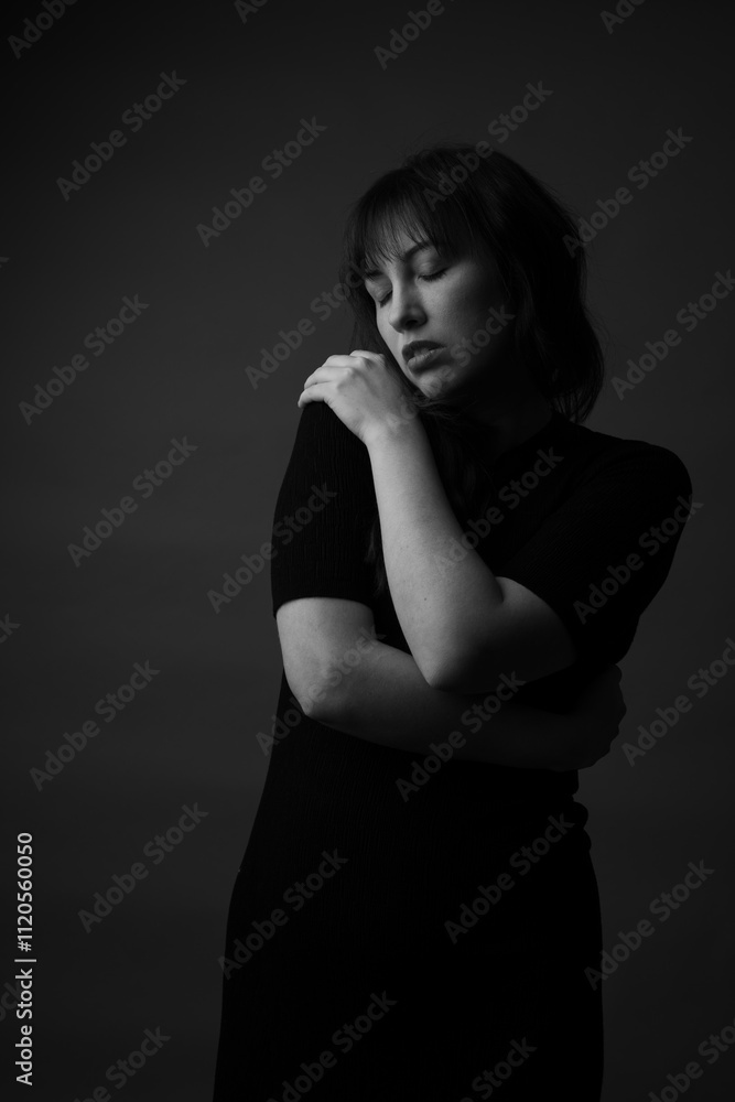Obraz premium Black and white dark portrait of a caucasian woman hugging herself with her eyes closed. She is wearing a black dress. This is a moody portrait. 