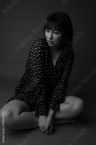 Black and white studio portrait of a caucasian woman with long dark hair with bangs. She is sitting on the floor cross legged wearing a long sleeve pleated dress. She is looking at the camera.