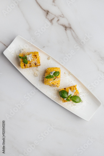 Three pieces of grilled corn on the cob are served on a white rectangular plate, topped with blue cheese, basil leaves, and grated cheese. Restaurant or cafe menu, cookbook recipe

