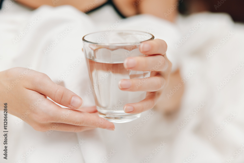 Woman holding glass of water sitting on bed with white comforter and pillows Woman holding glass of water sitting on bed with white comforter and pillows