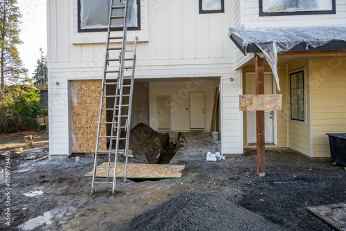 New house construction in progress, view into open garage with dirt floor and open plumbing pipe trench, extension ladders and newly installed interior doors
