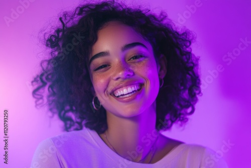 Joyful young woman with curly hair smiling brightly in vibrant purple lighting in a relaxed indoor setting