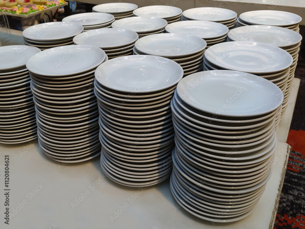 The image shows several stacks of white ceramic plates arranged neatly on a table. Each stack contains a large number of plates, indicating that this setup is likely for a large event or buffet The image shows several stacks of white ceramic plates arranged neatly on a table. Each stack contains a large number of plates, indicating that this setup is likely for a large event or buffet