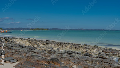 The rocky coast of the ocean. Boulders are scattered near the turquoise water. Boats at sea. There is a green tropical island in the distance. Mountains on the horizon. Clear  sky. Madagascar.  photo