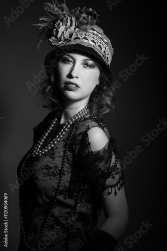 Black and white studio portrait of a caucasian woman dressed in 1920's fashion flapper outfit. She has shoulder length curly hair and is wearing feather hat. 