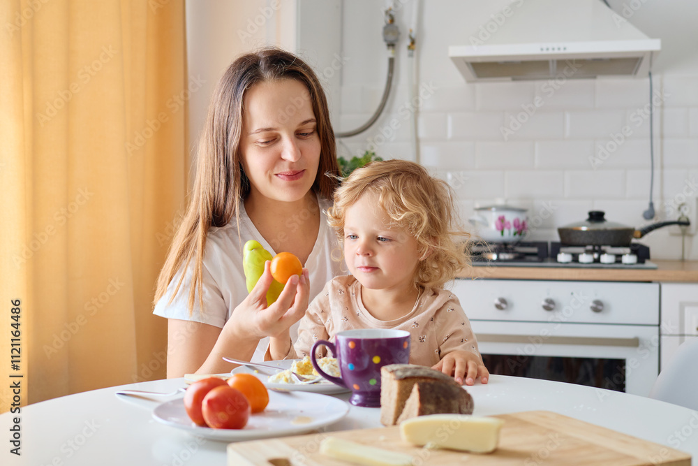 A mother shares fresh fruits and vegetables with her young child at a cozy kitchen table during breakfast. Warm sunlight fills the room, promoting a cheerful morning A mother shares fresh fruits and vegetables with her young child at a cozy kitchen table during breakfast. Warm sunlight fills the room, promoting a cheerful morning