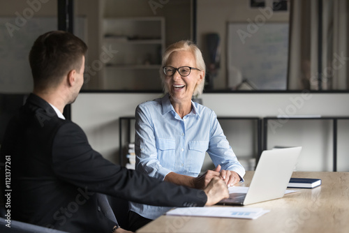 Two colleagues, aged woman and young man engaged in positive professional conversation met in company office with laptop, laugh, discuss positive outcome, project completion, business-related topics