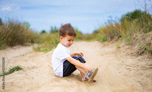 Child playing in sandy path on a sunny day. Young boy sits on a sandy trail, removing his shoe while enjoying a bright and playful outdoor moment.