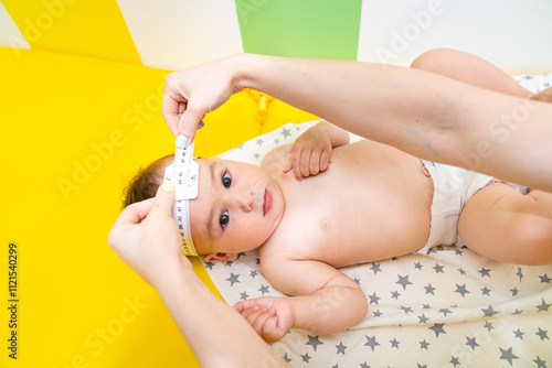 Baby health check with measuring tape indoors. A caregiver measures the head circumference of an infant lying on a colorful mat in a bright, cheerful room.