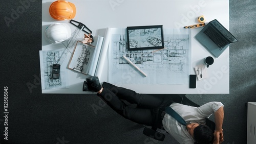 Top down view of relaxed architect engineer working on blueprint and putting leg on meeting table. Aerial view of skilled interior designer lie on the chair while sleeping in break time. Alimentation. photo