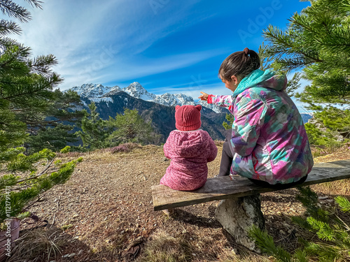 Mother with small child sitting on wooden bench on top of Monte Nebria Est with panoramic view of snow capped mountains of Julian Alps, Friuli Venezia Giulia, Italy. Family hike in alpine wilderness photo