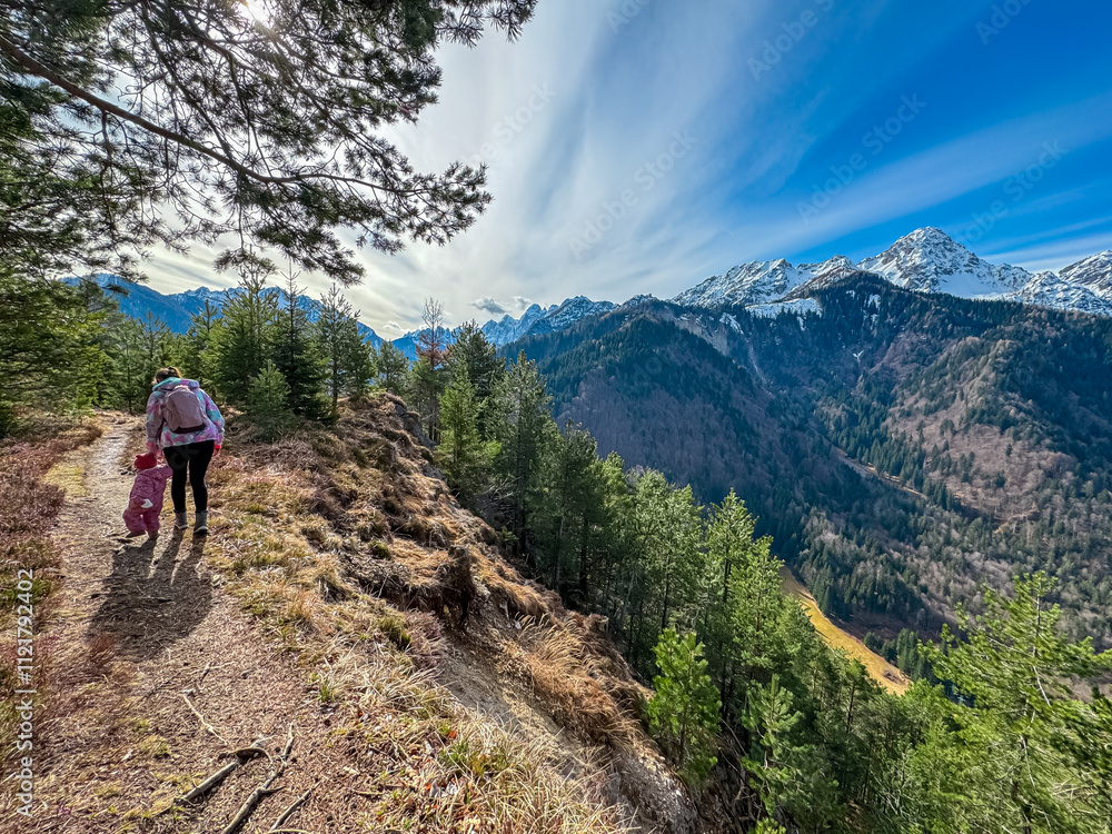 Mother walking with toddler on scenic hiking trail looking at snow capped mountain peaks of Julian Alps seen from top of Monte Nebria Cima Est, Friuli Venezia Giulia, Italy. Wanderlust in early spring Mother walking with toddler on scenic hiking trail looking at snow capped mountain peaks of Julian Alps seen from top of Monte Nebria Cima Est, Friuli Venezia Giulia, Italy. Wanderlust in early spring