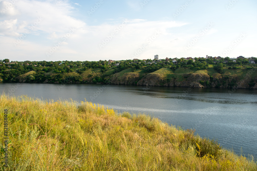 Scenic view of a tranquil river with lush green hills and a small village in the background under a clear blue sky. Perfect for nature, travel, and landscape themes Scenic view of a tranquil river with lush green hills and a small village in the background under a clear blue sky. Perfect for nature, travel, and landscape themes