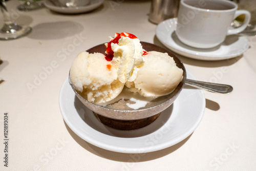A shiny metal bowl holds two scoops of creamy vanilla ice cream, crowned with whipped cream and drizzled in vibrant strawberry sauce, set atop a crisp white tablecloth with elegant tableware.