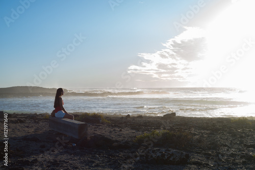 Mujer joven sentada en un banco junto al mar viendo la puesta de sol en un día con mar revuelto, al atardecer de un día de verano photo