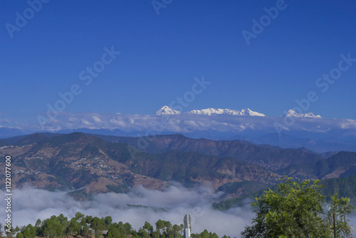 Snow covered Himalayan peaks including Mt.Trishul and Nanda Devi rise above a serene valley with green hills and dense clouds rolling over pine-covered slopes. View from a village in uttarakhand india photo