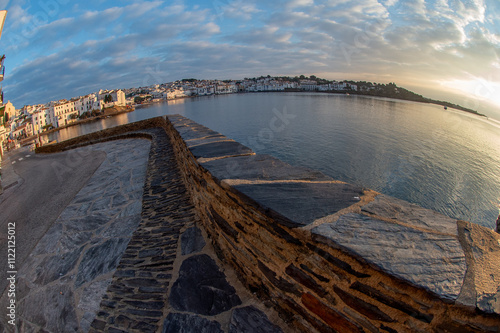 Wallpaper Mural vue originale du village de Cadaquès en Espagne, prise au grand angulaire avec une rue et une murette qui serpentent pour y aller, et la mer légèrement courbée le soleil se lève au fond sur le côté Torontodigital.ca