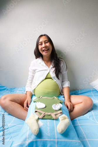 pregnant woman sitting on bed with joyful expression, holding baby clothes and accessories on her belly, symbolizing excitement and preparation for motherhood