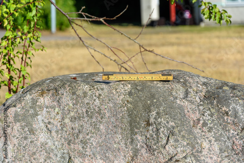 A ruler rests on a large rock in a park, surrounded by grass and trees. The sun shines brightly, creating shadows and highlighting the texture of the stone