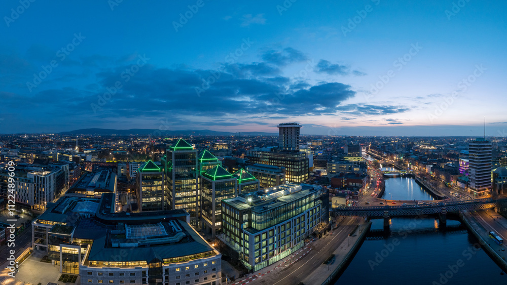 Fototapeta premium A dusk view of Dublin's modern skyline with the iconic green-lit Bank of Ireland building, a river reflecting the city lights, and a bridge crossing over it.