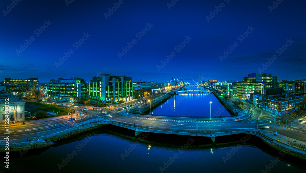 Fototapeta premium A dusk view of Dublin's modern skyline with the iconic green-lit Bank of Ireland building, a river reflecting the city lights, and a bridge crossing over it.
