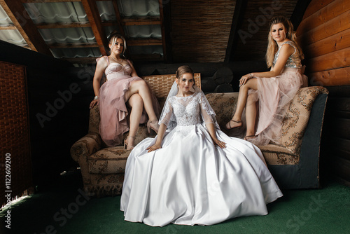 On either side of the bride sit two bridesmaids in soft pink dresses. One of them sits on the left, the other on the right. They are all located in a room with wooden walls and a roof with visible bea photo