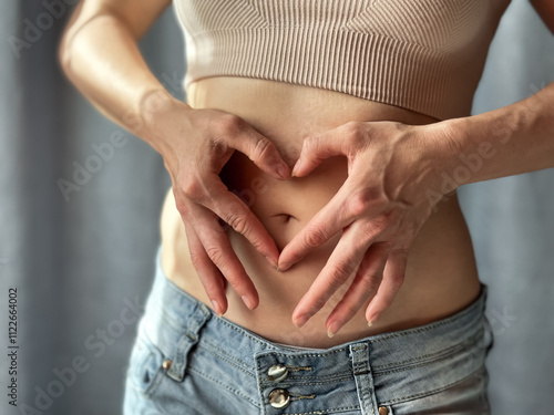 Close up of slim woman in short top and jeans forming heart shape with her hands on her bare stomach. Concept of body positivity, self love, and health awareness. Lifestyle photo. photo