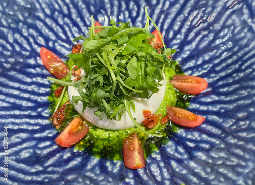 a refreshing serving of burrata cheese with cherry tomatoes, pesto sauce and rocket salad on a blue and white ceramic plate photo