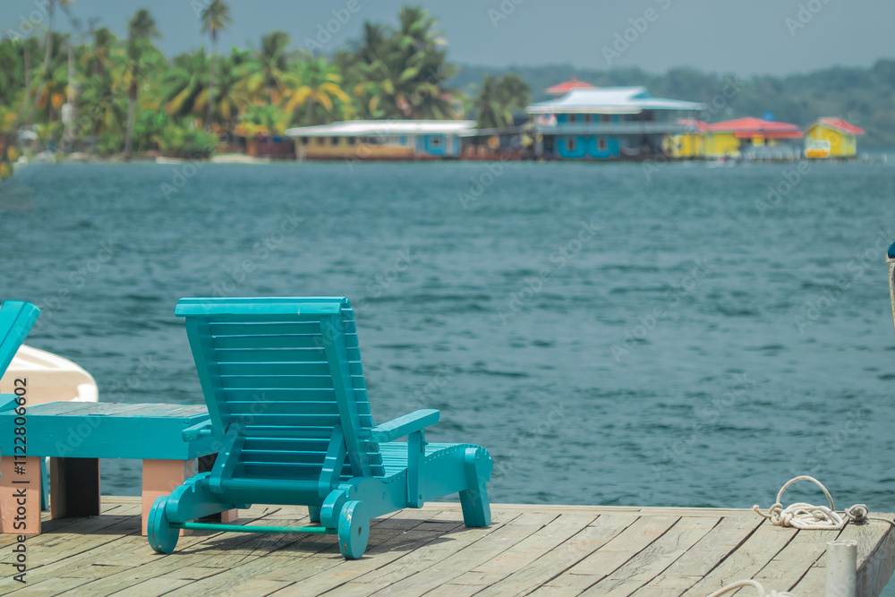 Beach chairs for resting at a pier or dock at beautiful destination of Bocas del Toro, Panama. Visible colorful houses, water and palms in the background.