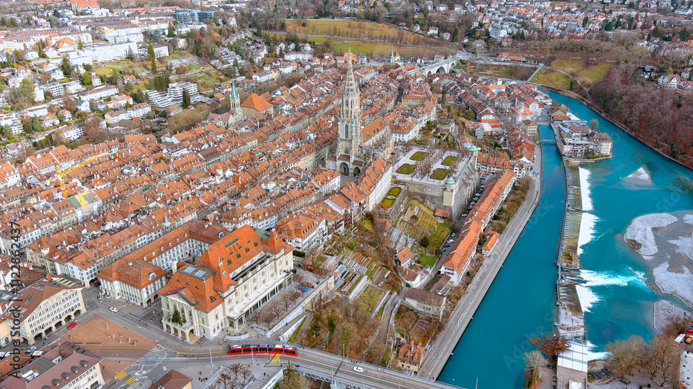 Fototapeta premium Breathtaking aerial image of Bern’s Old Town featuring the historic Münster Cathedral, red-roofed houses, and the serene Aare River. A captivating mix of Swiss heritage and natural beauty.