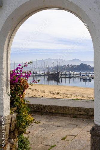 Museum caravel La Pinta in the fishing port of Baiona seen through an arch with purple flowers. Pontevedra, Galicia, Spain. photo