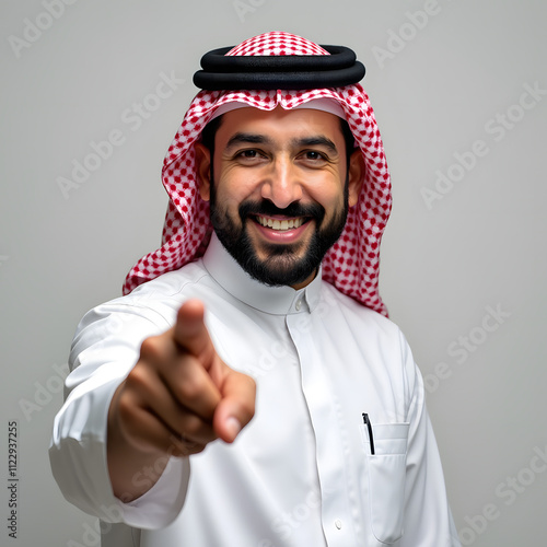 Pointing to something, looking at the camera with gestures of happiness and pleasure, portrait of a smiling Saudi Gulf Arab man wearing a traditional thobe and shemagh pointing with his index finger photo