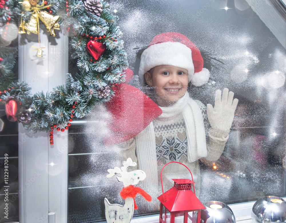 little girl in santa hat and with gifts shines a lantern into the window from winter street