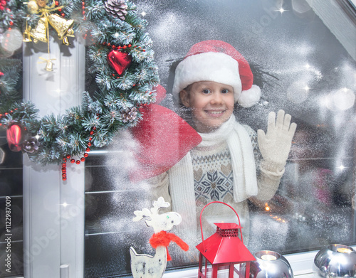 Obraz na płótnie little girl in santa hat and with gifts shines a lantern into the window from winter street w salonie