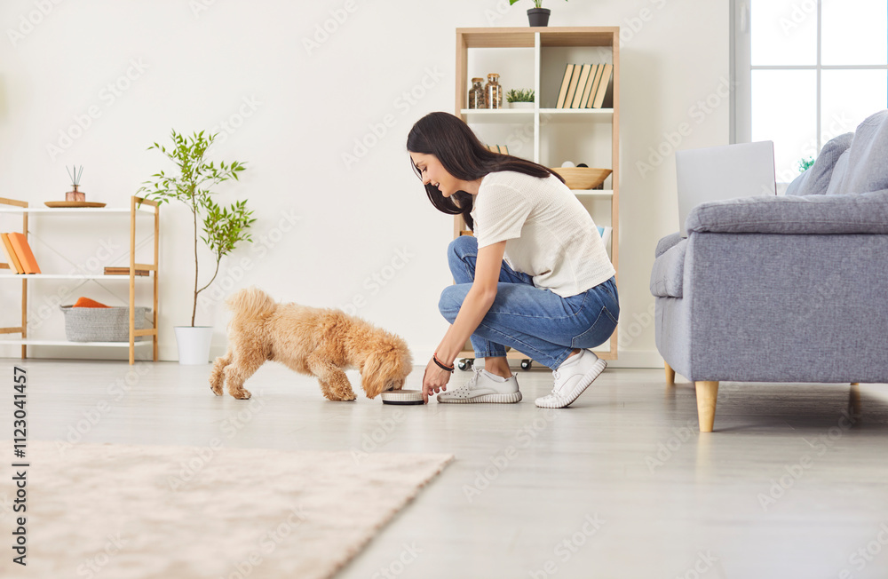 © Studio Romantic - Positive woman feeding dog from a bowl with pet food at home. The domestic pet happily eats, showcasing the bond between the owner and animal friend in a peaceful home environment.