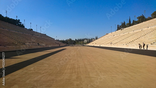 Athens, Greece - 29.3.2018: Tourists walking along the track on the field of Panathenaic Stadium with marble steps on both sides and flags at the end under a clear blue sky in the summer before Covid