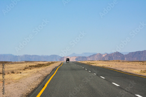 A flat road in the desert in perspective with a car driving in the distance. Tourist trip by own transport. Small mountains in the distance