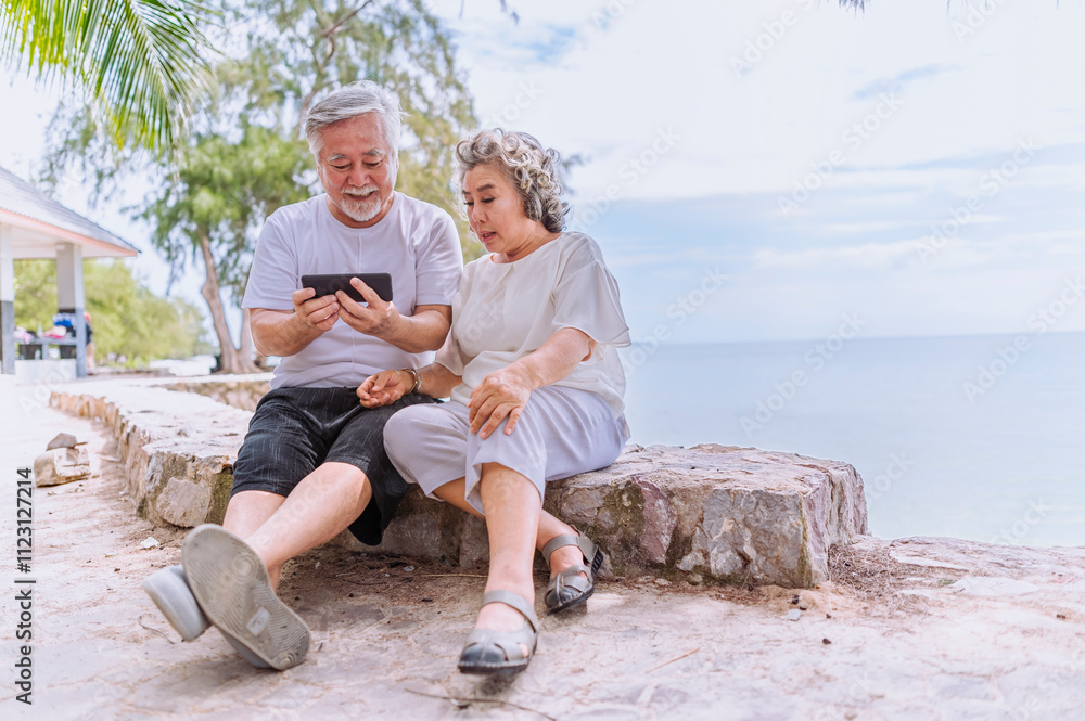 Happy asian senior retired couple; relax elder man and woman enjoying with smartphone apps, middle-aged wife enjoy online entertainments in retired vacation at sea beach outdoor.