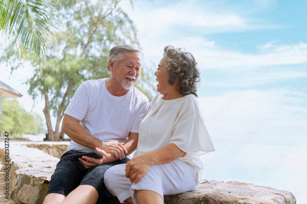 Happy asian senior retired couple; relax elder man and woman enjoying with smartphone apps, middle-aged wife enjoy online entertainments in retired vacation at sea beach outdoor.