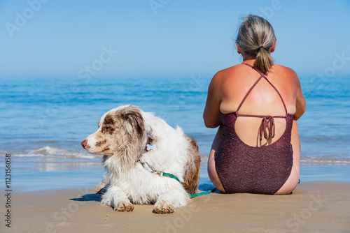 A woman with her back turned sitting on a beach with her pet dog with the sea in the background photo