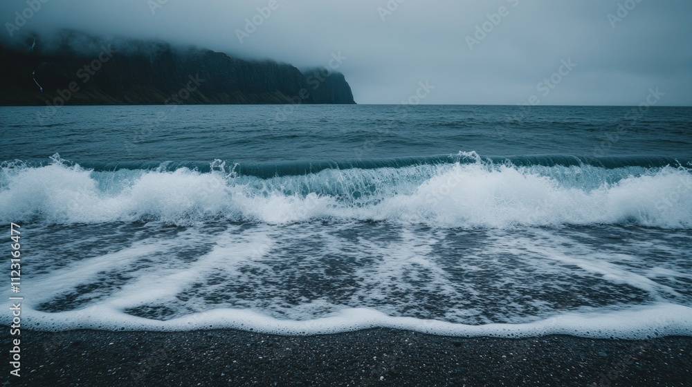 Obraz premium Foamy waves crashing on a dark beach, with a misty mountain range in the distance.