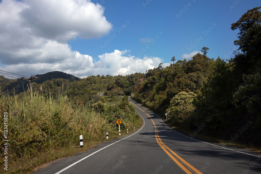 Obraz premium Scenic winding road through lush greenery and mountains under a blue sky with fluffy clouds.