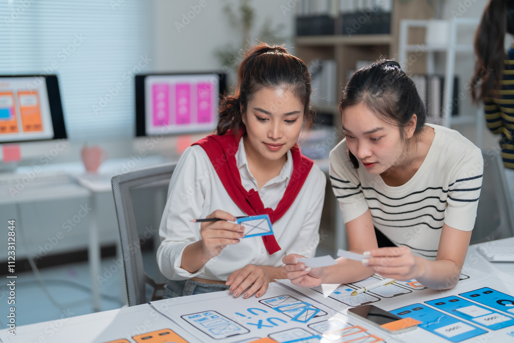 Fototapeta premium Two female ux designers are analyzing mobile app wireframes and discussing user interface improvements in a modern office environment, collaborating on enhancing user experience